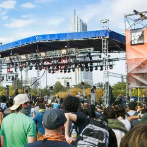 A large daytime crowd is gathered in front of a massive stage with trussing and lights covered with a blue tarp with a pillar on the side draped with a vinyl banner that says Fun Fun Fun Fest.