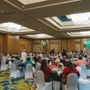An assorted group of people sit banquet style inside an elegant ballroom.