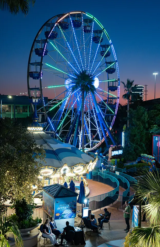 A large Ferris wheel, bright lights line its spokes as it juts into a skyline with the fading pink glow of the evening as nightfall descends while a group of people sit nearby a carousel in the foreground.